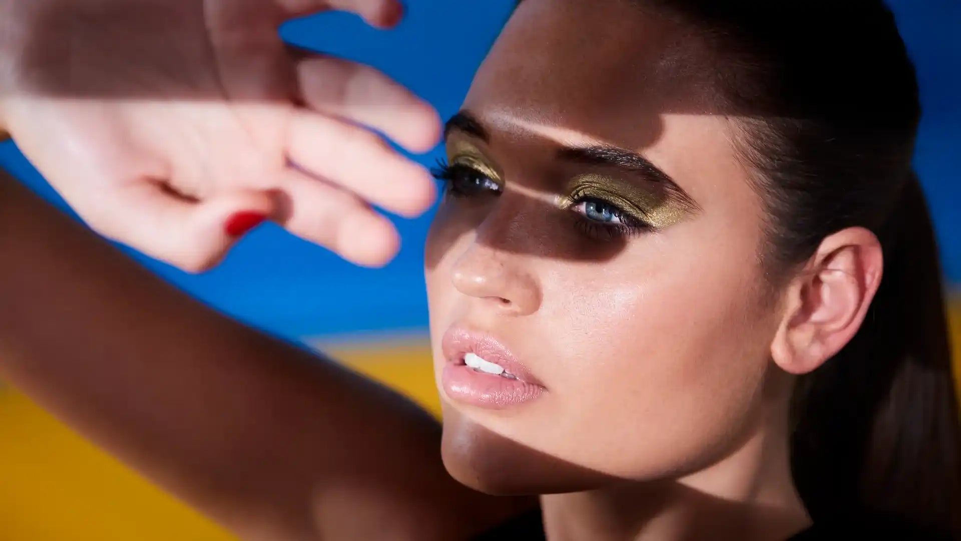 A woman’s face with dramatic eye makeup and a hand shading her eyes.