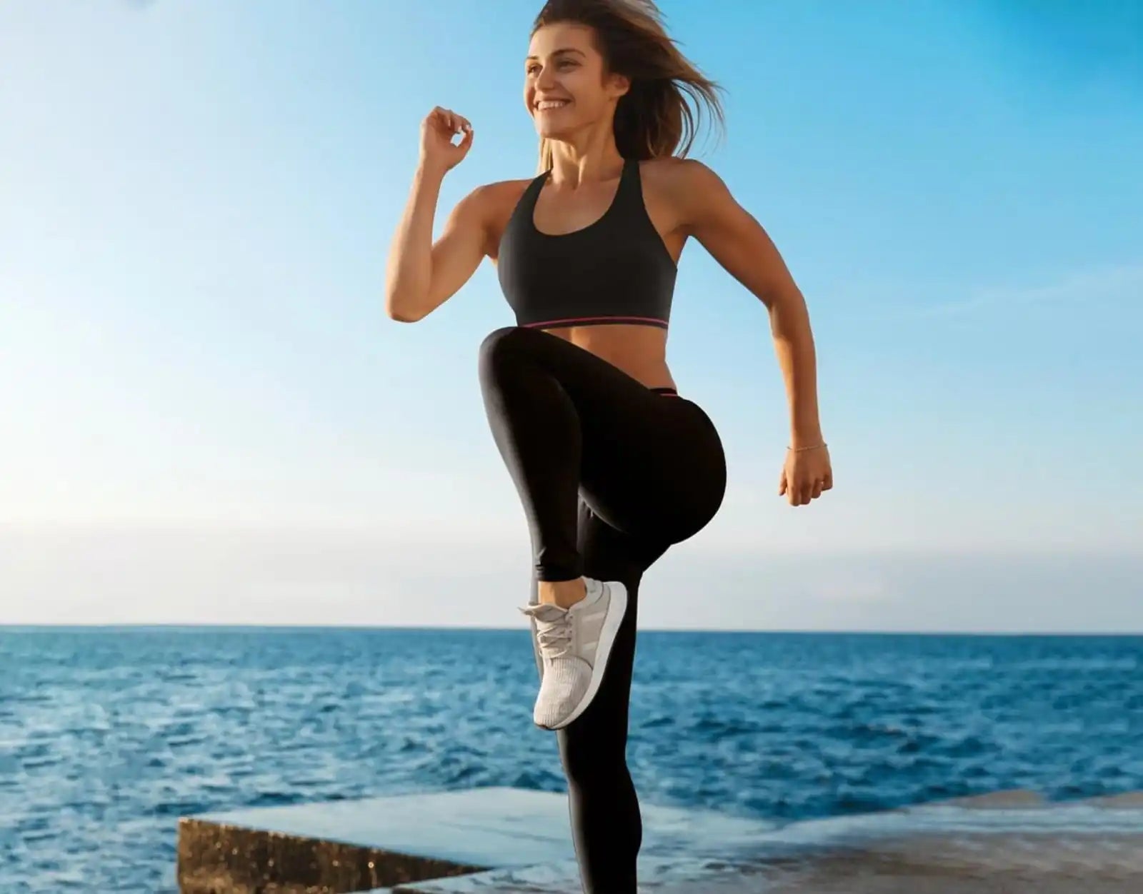 A smiling woman in a black sports bra and leggings, mid-stride in white athletic shoes.