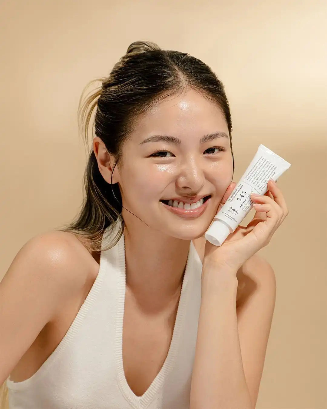 A young woman with glowing skin, holding a white tube of skincare product.