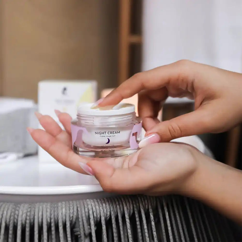 A jar of night cream, with a light purple label, being held by two hands, one of which is dipping a finger into the cream.