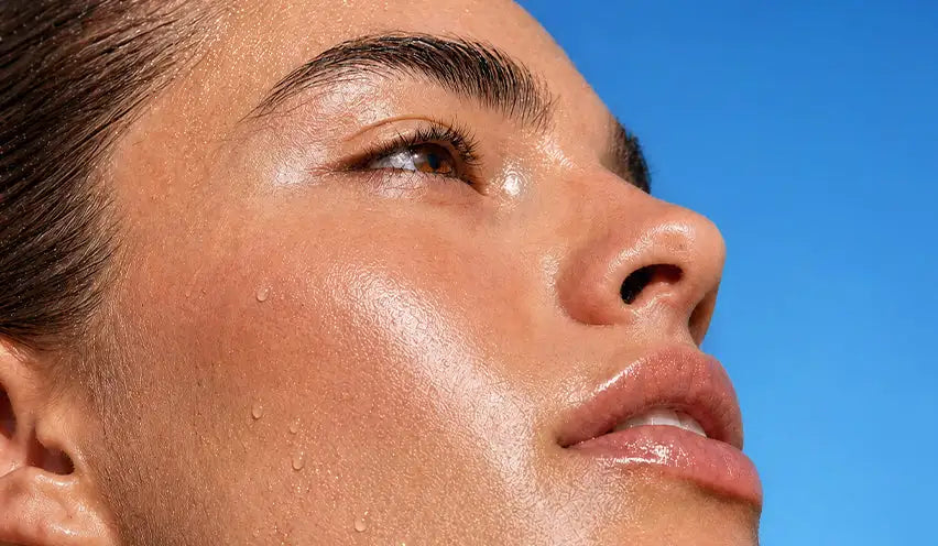A close-up of a woman’s face with glistening skin and visible sweat droplets against a bright blue sky.