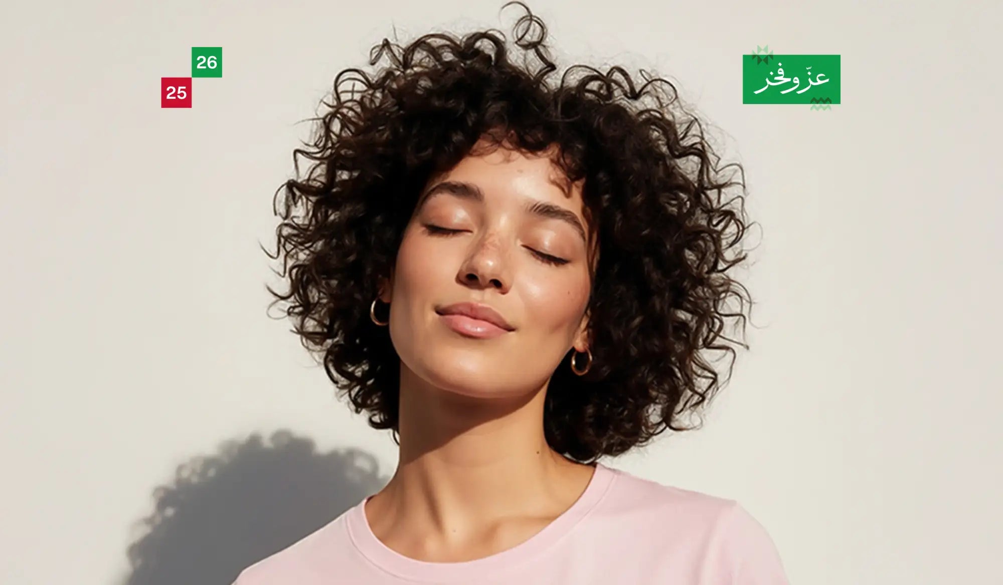 A woman with voluminous dark curly hair wearing a soft pink crew-neck top.