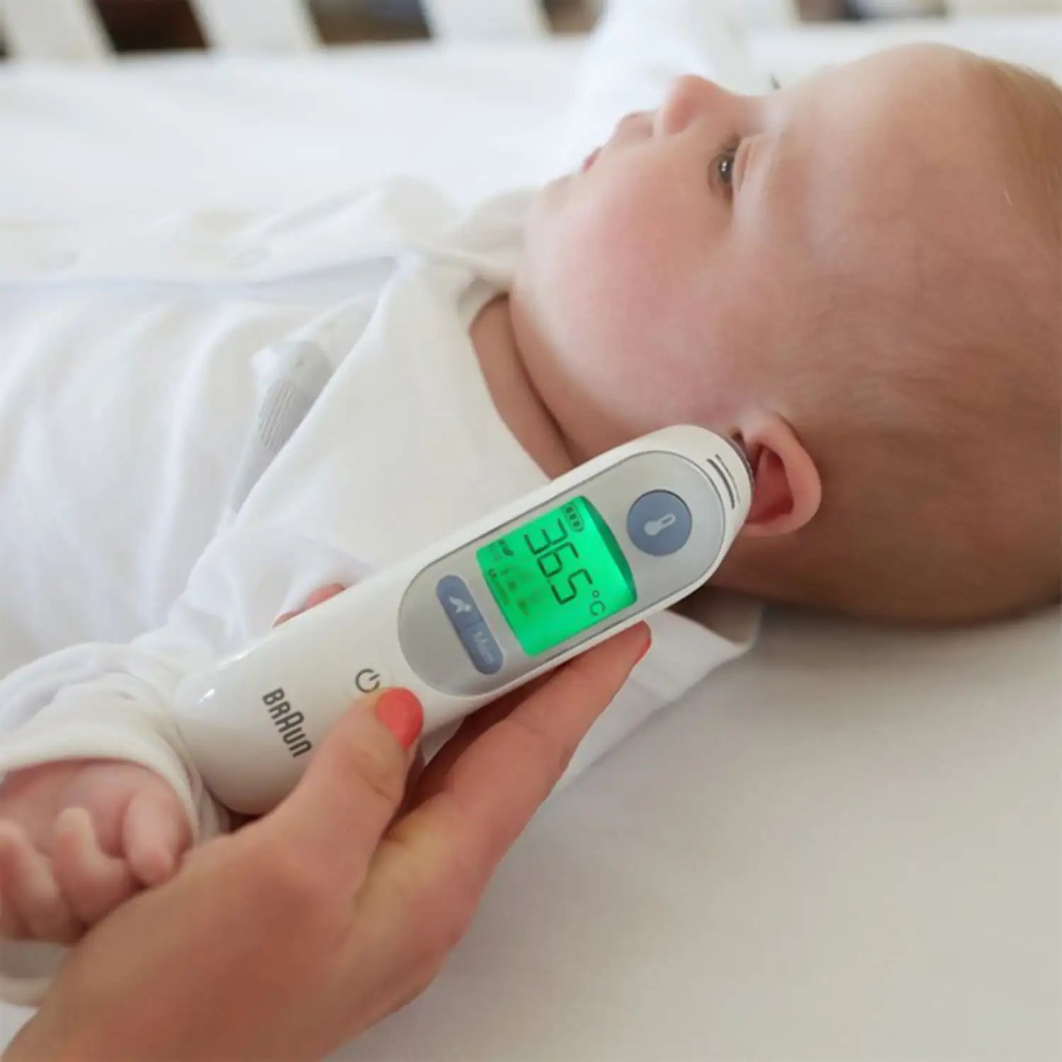 White braun digital infrared thermometer with a green display showing 35.2°c being held near a baby’s ear.