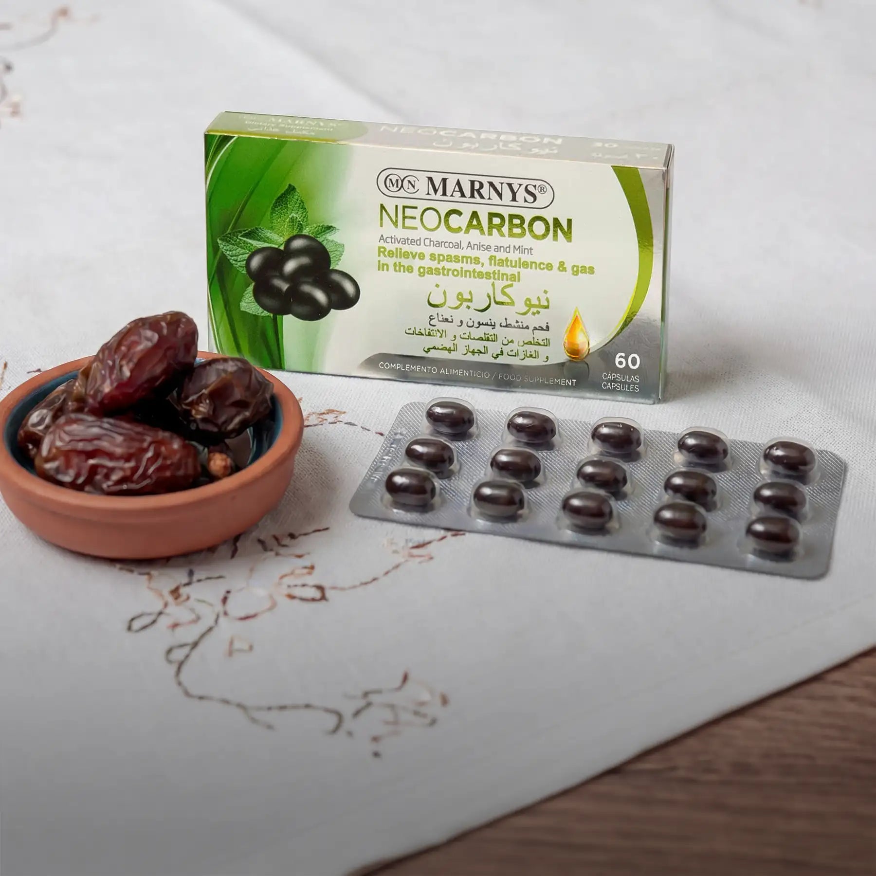 Green and white box of marnys neocarbon activated charcoal capsules with dark brown, glossy gelatin capsules arranged in a blister pack beside a small terracotta bowl filled with dates.