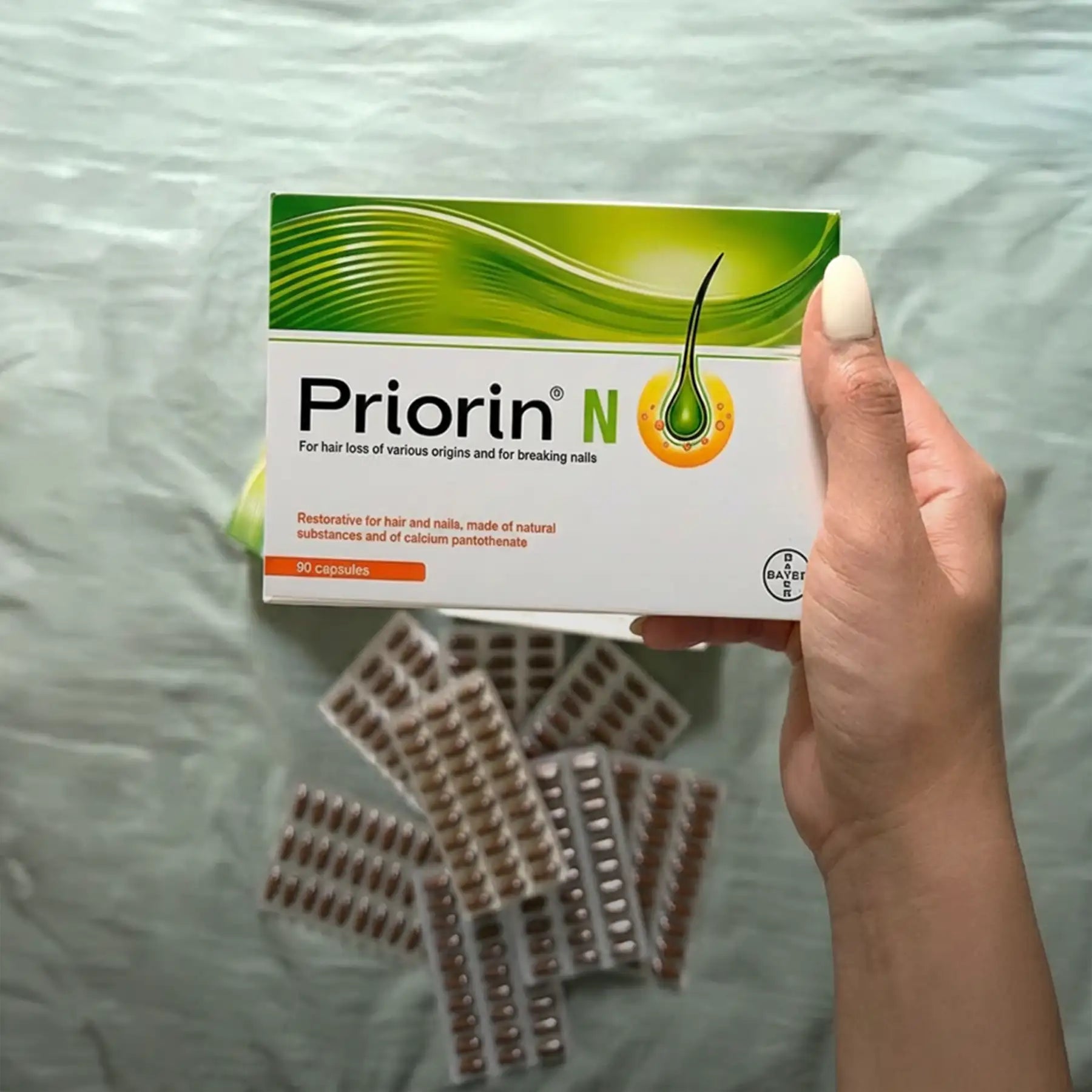 A hand holds a white and green box of priorin n, featuring an illustration of a hair strand with a drop of liquid beneath it.
