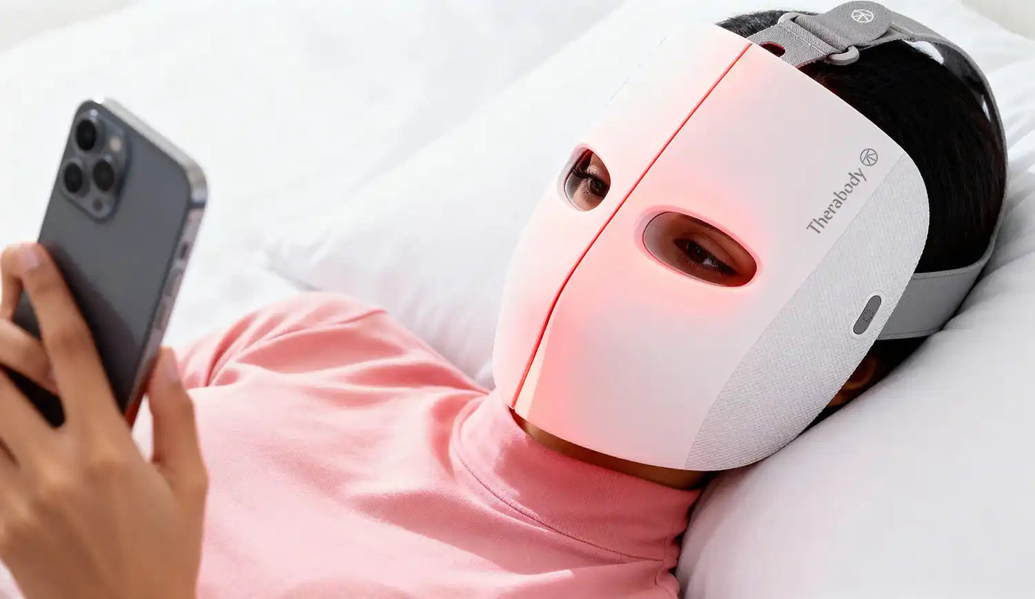 A person wearing a white LED light therapy mask and holding a smartphone.